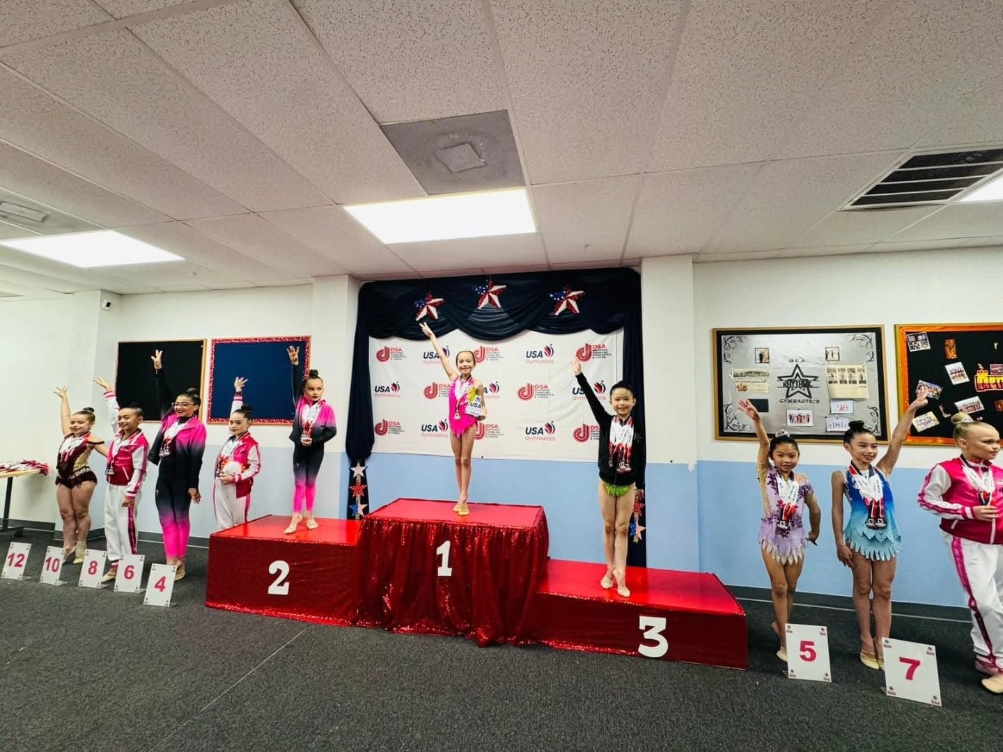 Young medalists wave from the awards podium in front of a USA Gymnastics backdrop