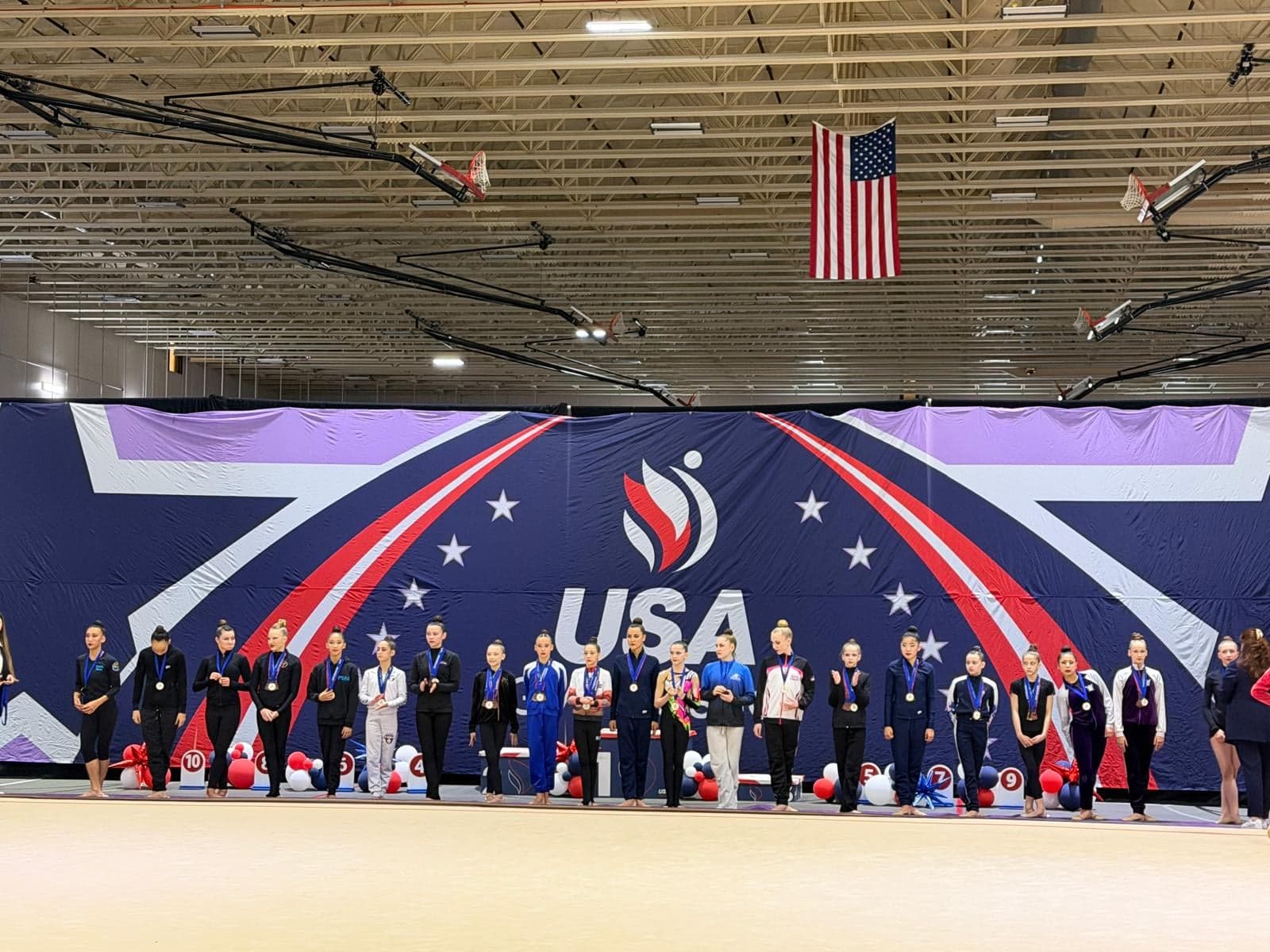 Finalists lined up on the awards podium under the American flag at the 2026 USAG Rhythmic Elite Qualifier