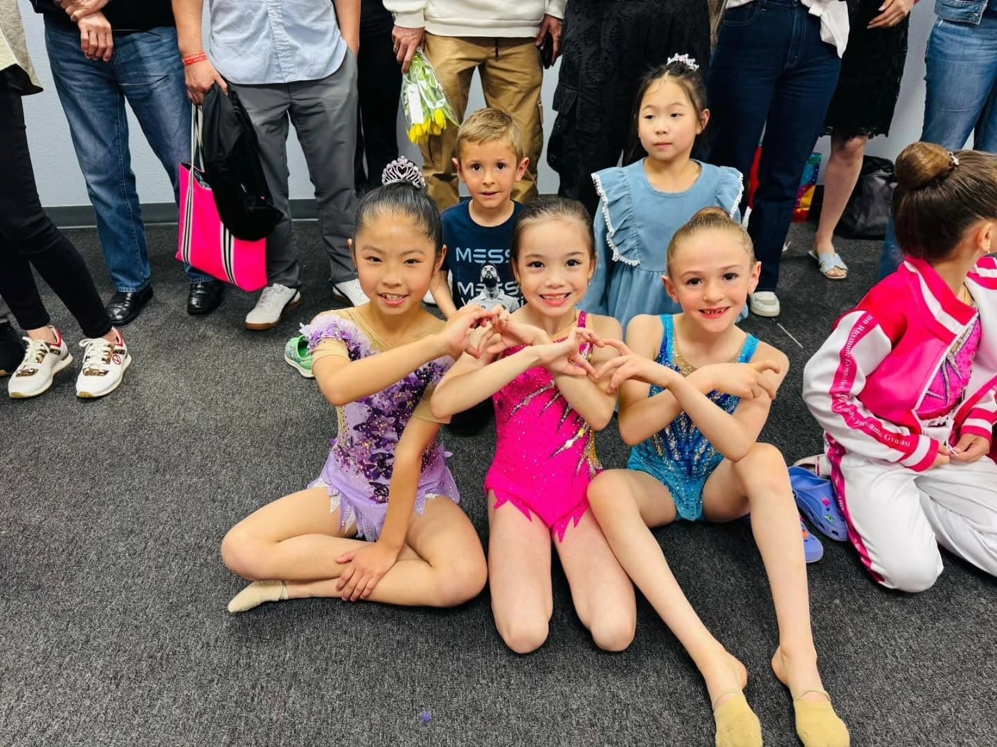 Three young gymnasts in competition leotards make heart hands together at a meet