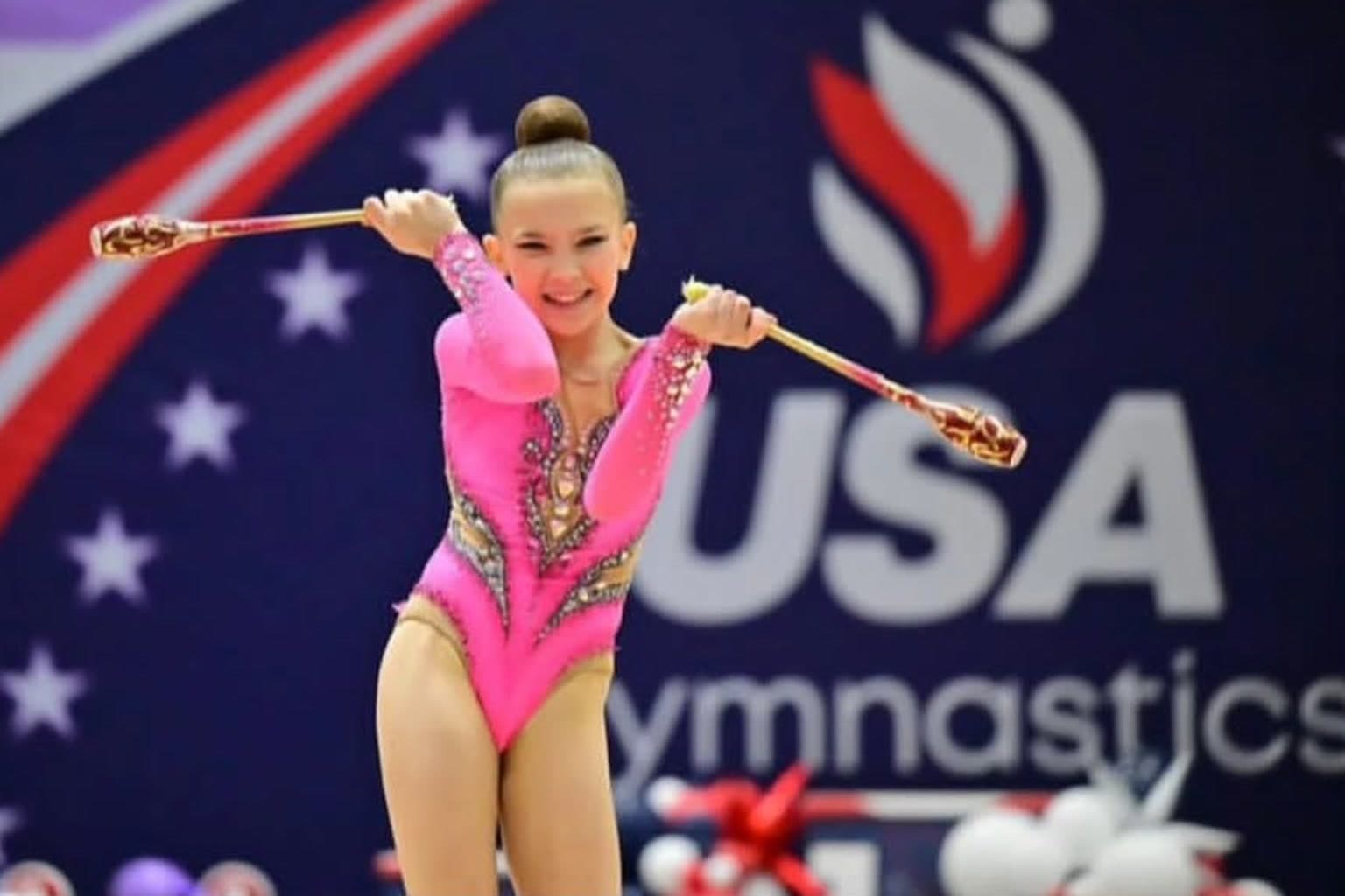 Rhythmic gymnast performing with clubs in front of the USA Gymnastics backdrop at a competition