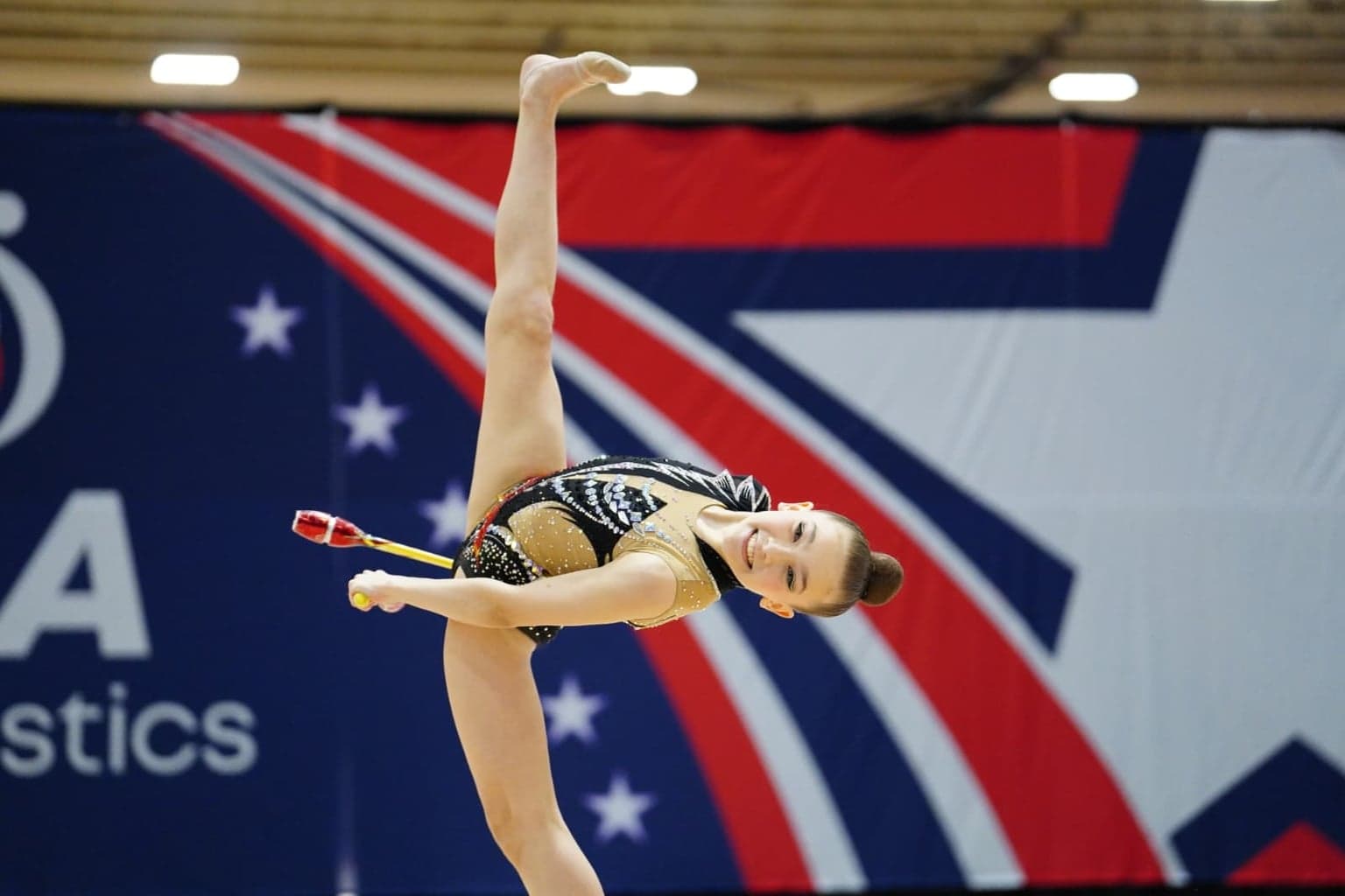 Rhythmic gymnast smiling while performing a balance skill with clubs during a competition routine