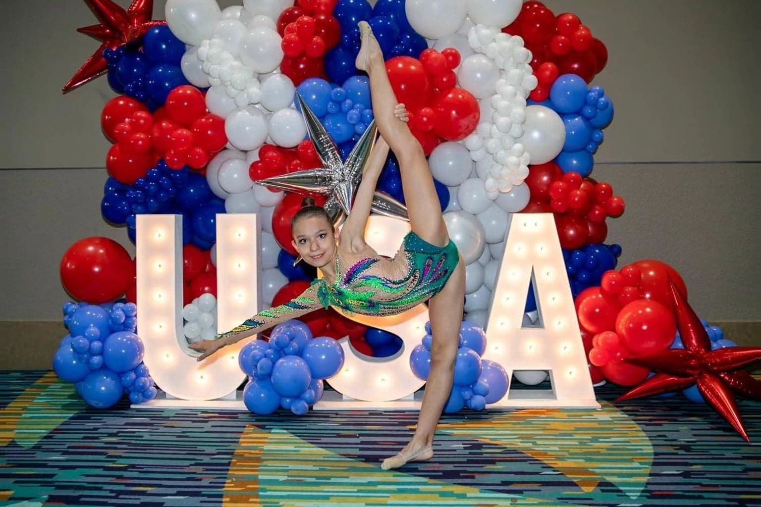 Rhythmic gymnast performing a standing split in front of red, white, and blue balloons and illuminated USA letters