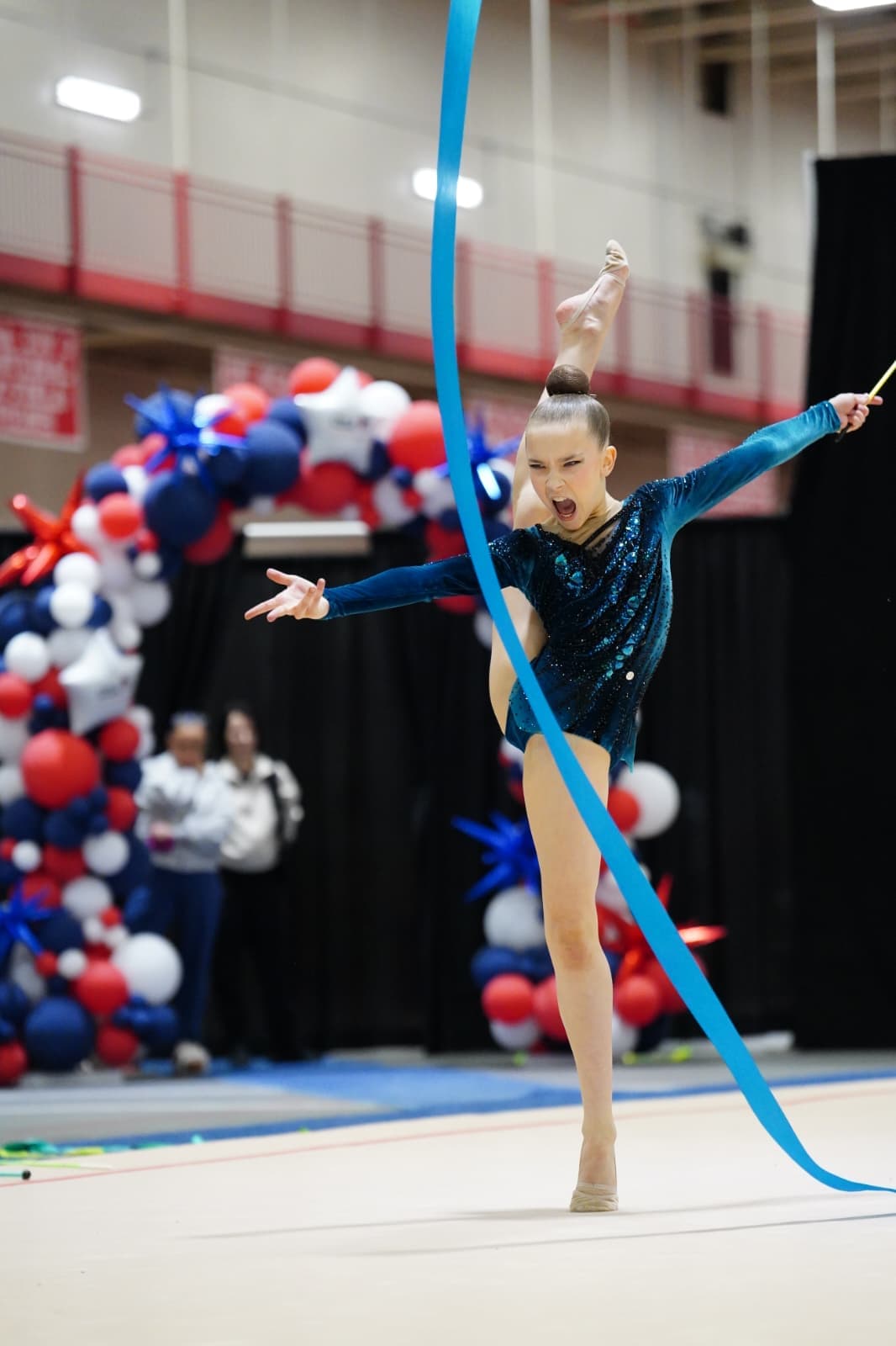 Rhythmic gymnast performing an expressive balance skill with a blue ribbon during competition.