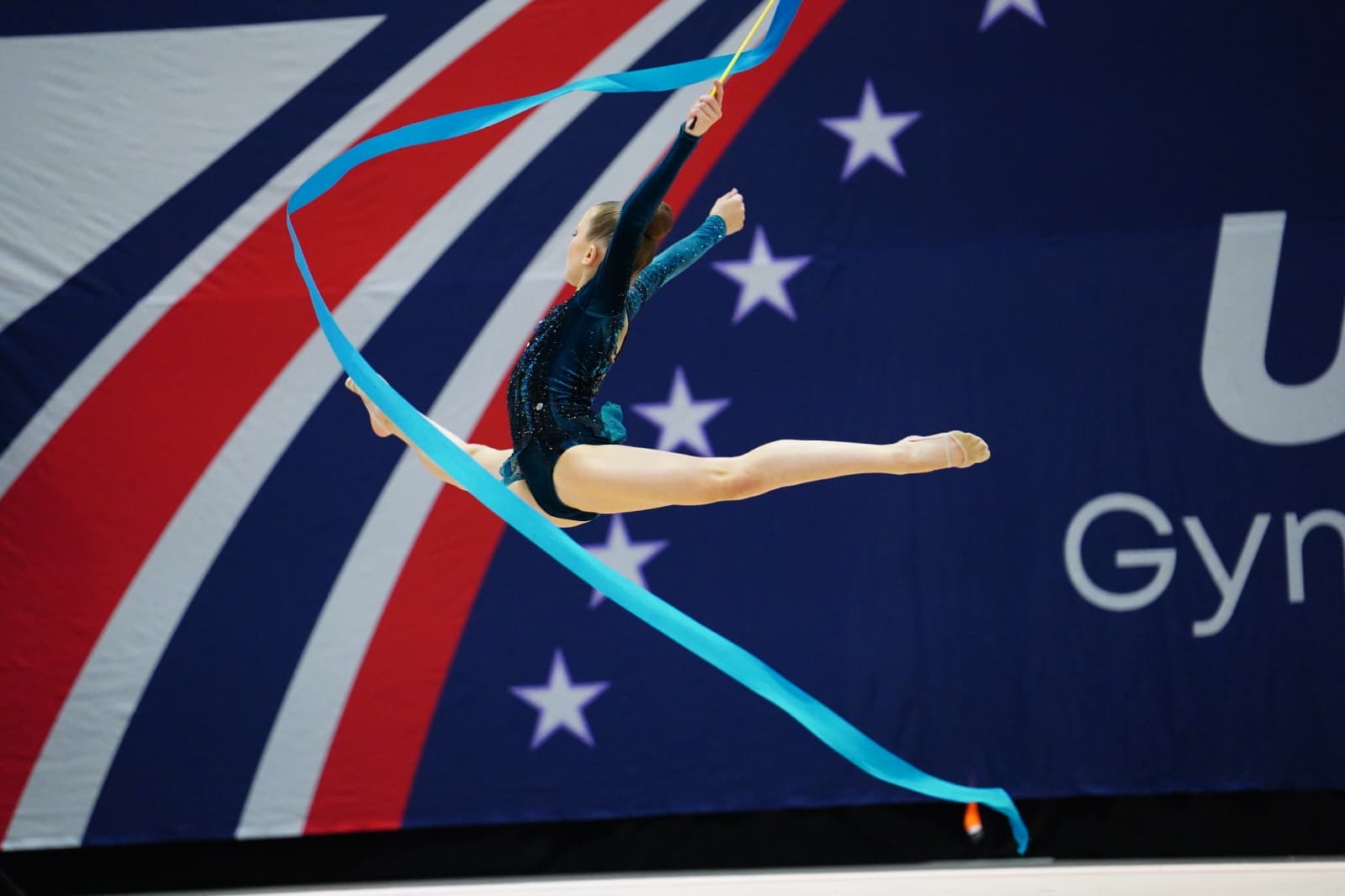 Rhythmic gymnast performing a split leap with a blue ribbon during competition.