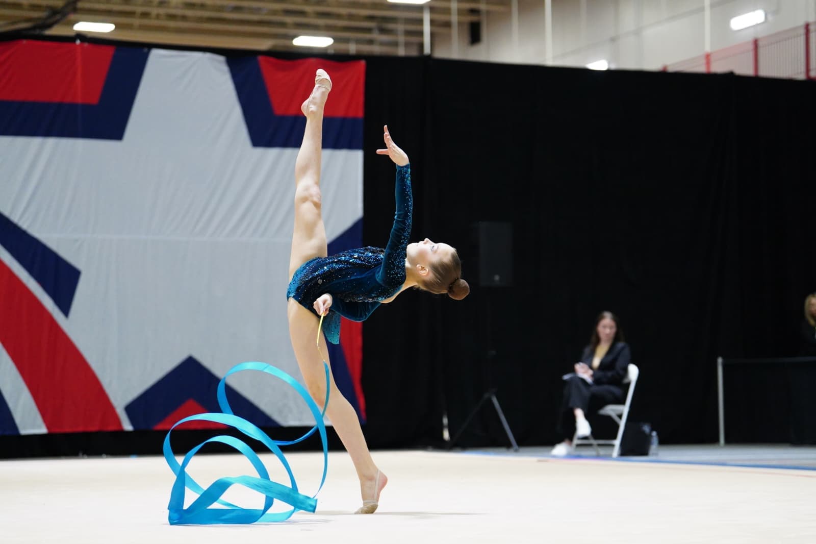 Rhythmic gymnast performing a high balance skill with a blue ribbon during competition.