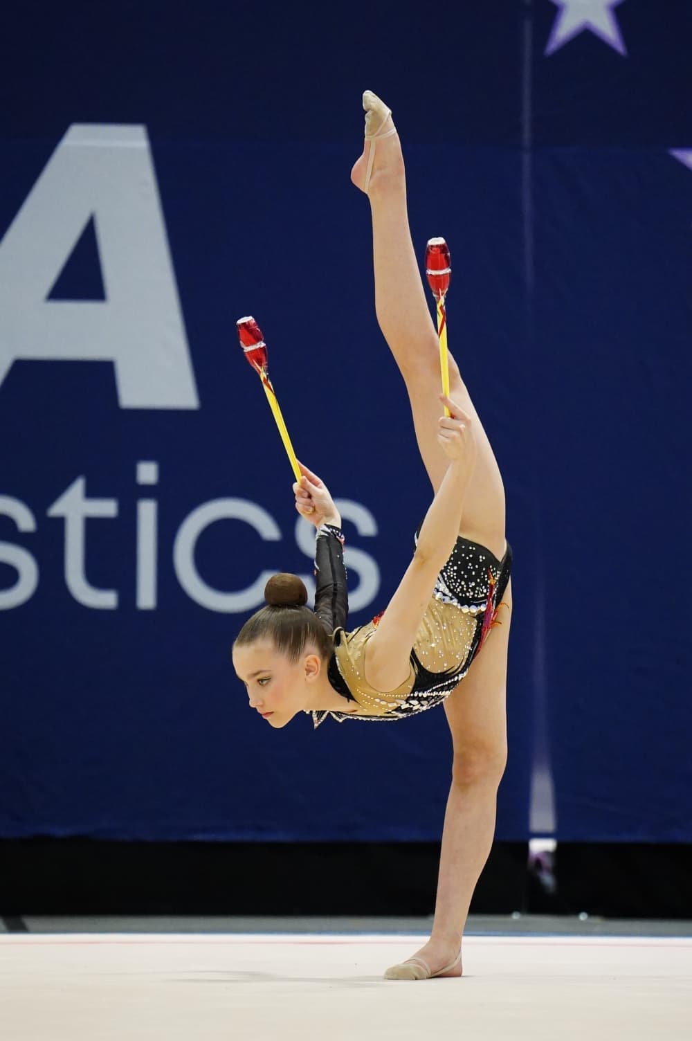 Rhythmic gymnast performing a high balance skill with clubs during a competition routine.