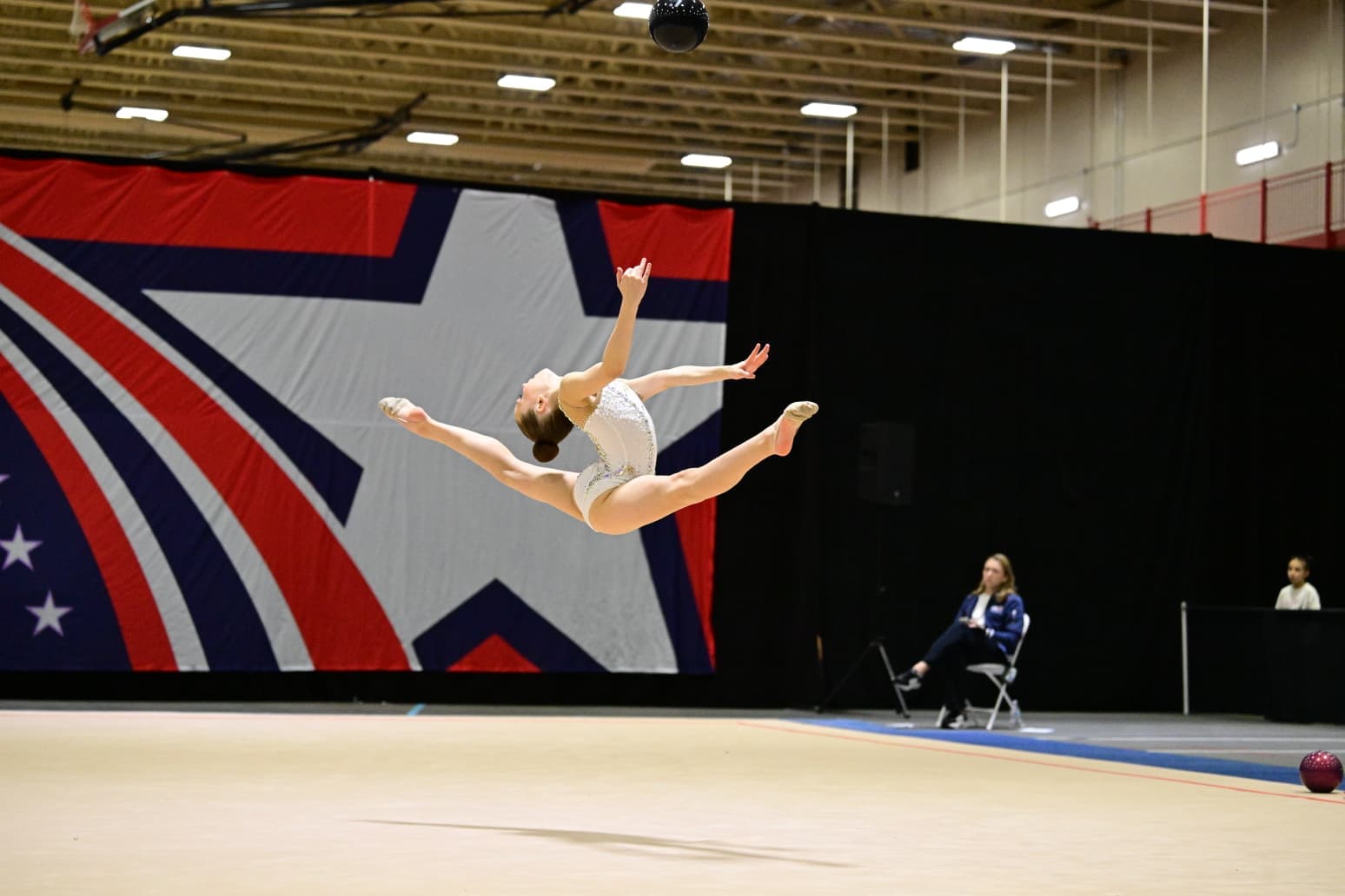 A rhythmic gymnast performs a powerful split leap across the competition floor, showing strength, flexibility, control, and artistry.